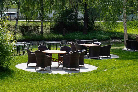 Outdoor dining area with brown rattan chair and tables surrounded by green grass and trees.