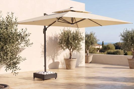 A large beige patio umbrella with a black central pole and square base, set on a sunlit stone terrace beside a cream-colored building with potted olive trees under a clear blue sky.