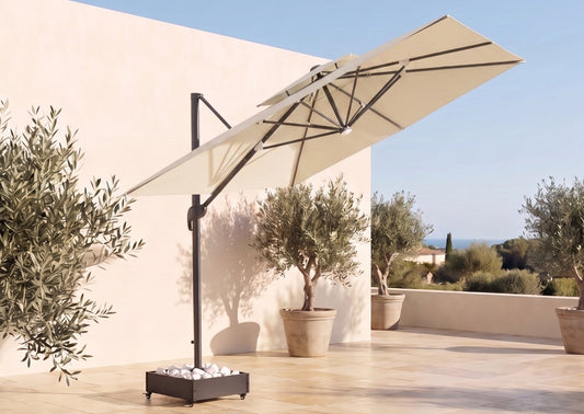 A large beige cantilever patio umbrella with a black metal frame and base, set on a sunlit stone terrace with potted olive trees and a beige stucco wall in the background.